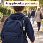 Dziecko widziane od tyłu idzie chodnikiem do szkoły, na szelce plecaka czarny brelok, w tle dorośli